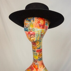 Puede incluir: Un sombrero fedora de fieltro negro con un ala ancha y una copa plana. El sombrero est&aacute; colocado sobre una cabeza de maniqu&iacute; con un dise&ntilde;o estampado de colores.