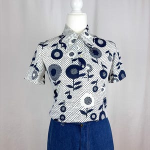 May include: A short-sleeved, collared shirt with a white background and a repeating pattern of navy blue floral and dot designs. The shirt is displayed on a mannequin and paired with dark blue denim pants.