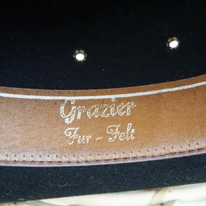 Puede incluir: Un sombrero de fieltro negro con una banda de cuero marr&oacute;n. La banda tiene el texto "Grazier Fur - Felt" grabado en letras plateadas.