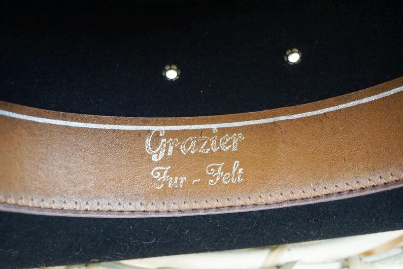 Puede incluir: Un sombrero de fieltro negro con una banda de cuero marr&oacute;n. La banda tiene el texto "Grazier Fur - Felt" grabado en letras plateadas.