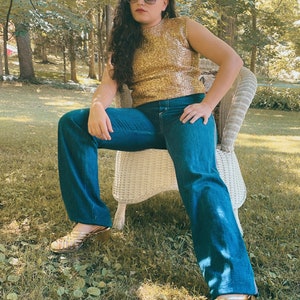 May include: A woman wearing a gold sequined top, blue jeans, and gold sandals sits on a white wicker chair in a grassy area.