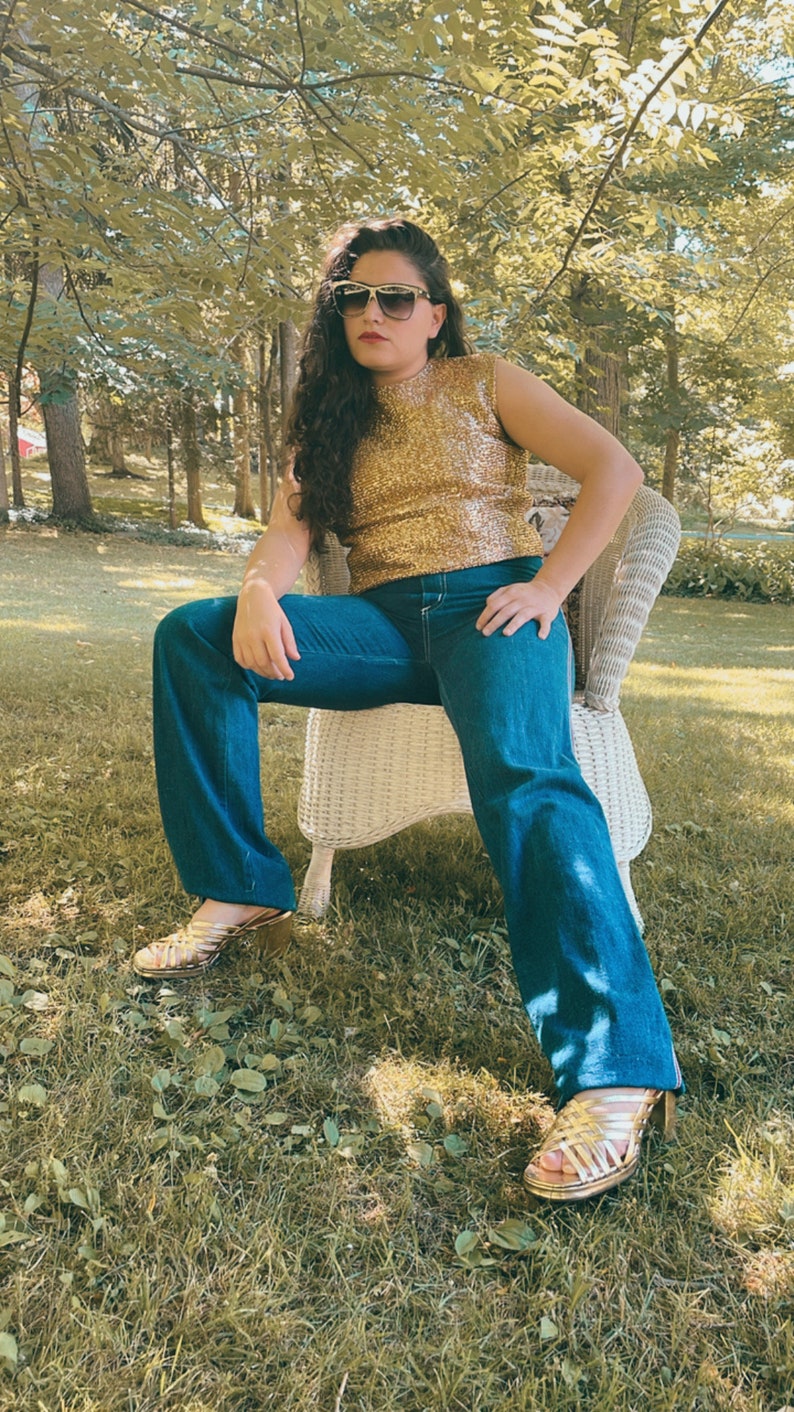 May include: A woman wearing a gold sequined top, blue jeans, and gold sandals sits on a white wicker chair in a grassy area.