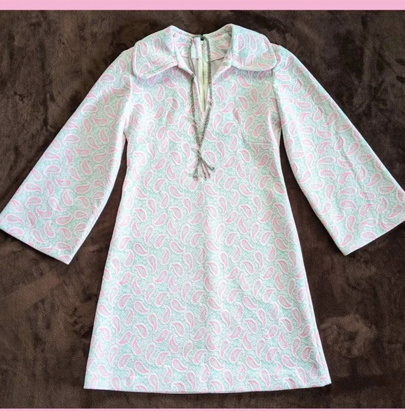 May include: A white and pink paisley patterned dress with a long collar and long sleeves. The dress has a V-neckline and a silver chain necklace.