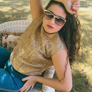 May include: A woman wearing a gold sequined top and blue jeans sits on a white wicker chair. She is wearing sunglasses and has her hair pulled back.