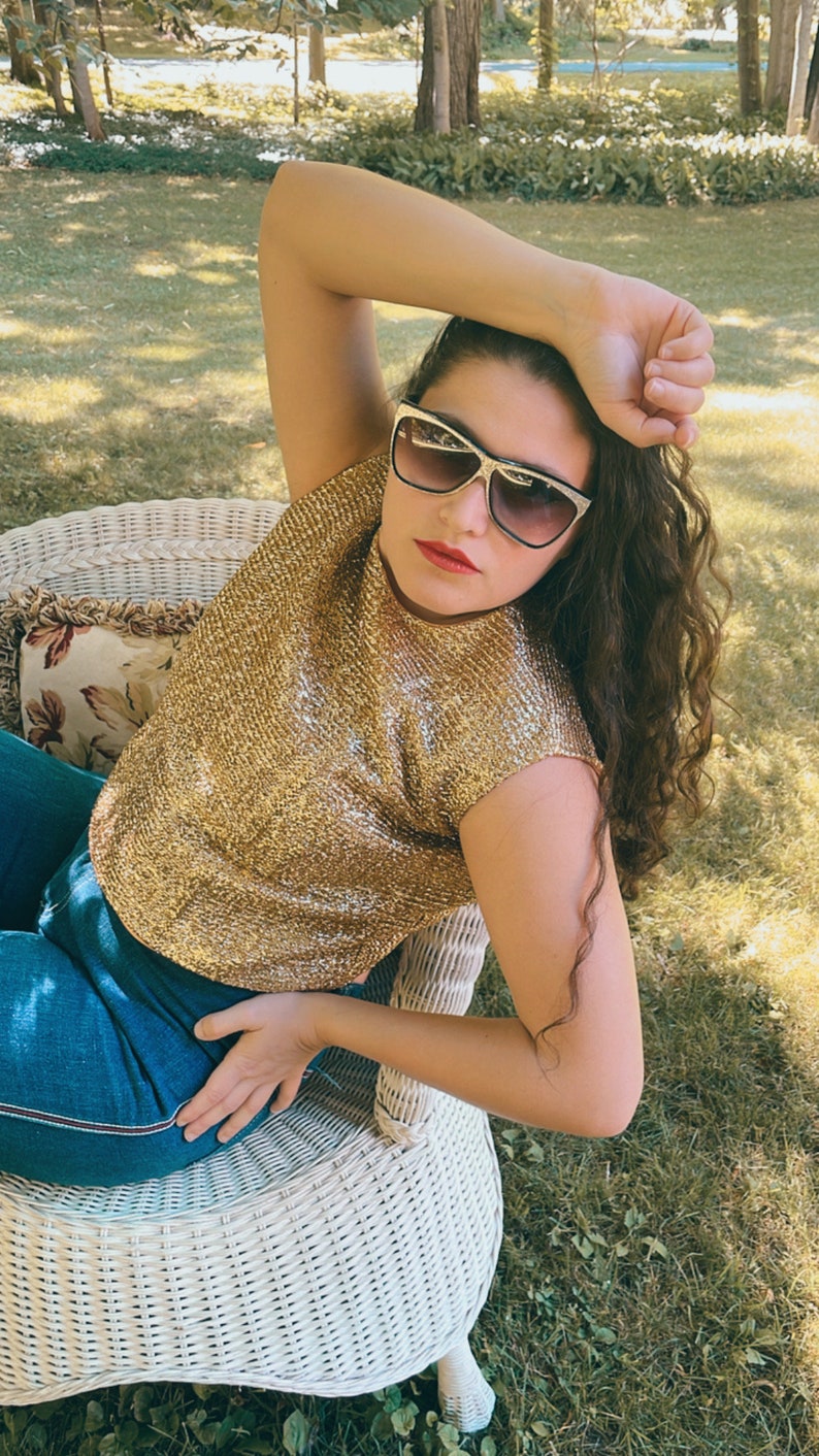 May include: A woman wearing a gold sequined top and blue jeans sits on a white wicker chair. She is wearing sunglasses and has her hair pulled back.