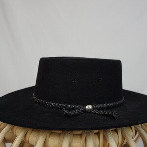 Puede incluir: Un sombrero fedora de fieltro negro con una banda de cuero trenzada y un bot&oacute;n plateado.