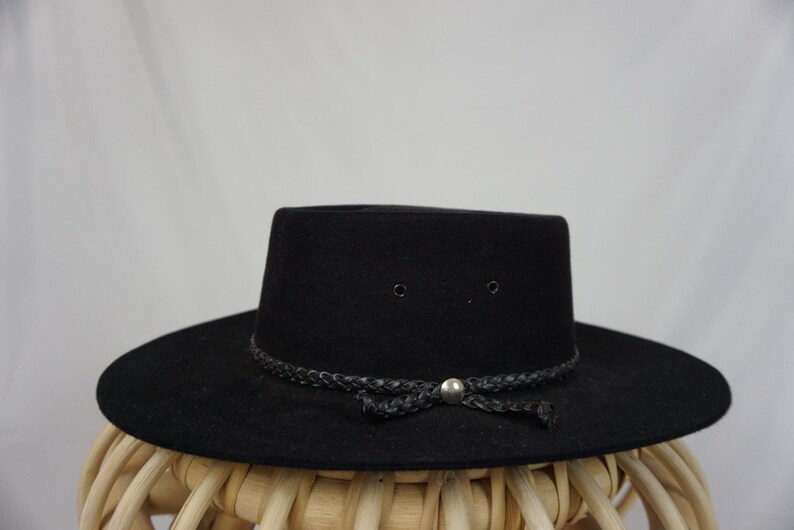 Puede incluir: Un sombrero fedora de fieltro negro con una banda de cuero trenzada y un bot&oacute;n plateado.