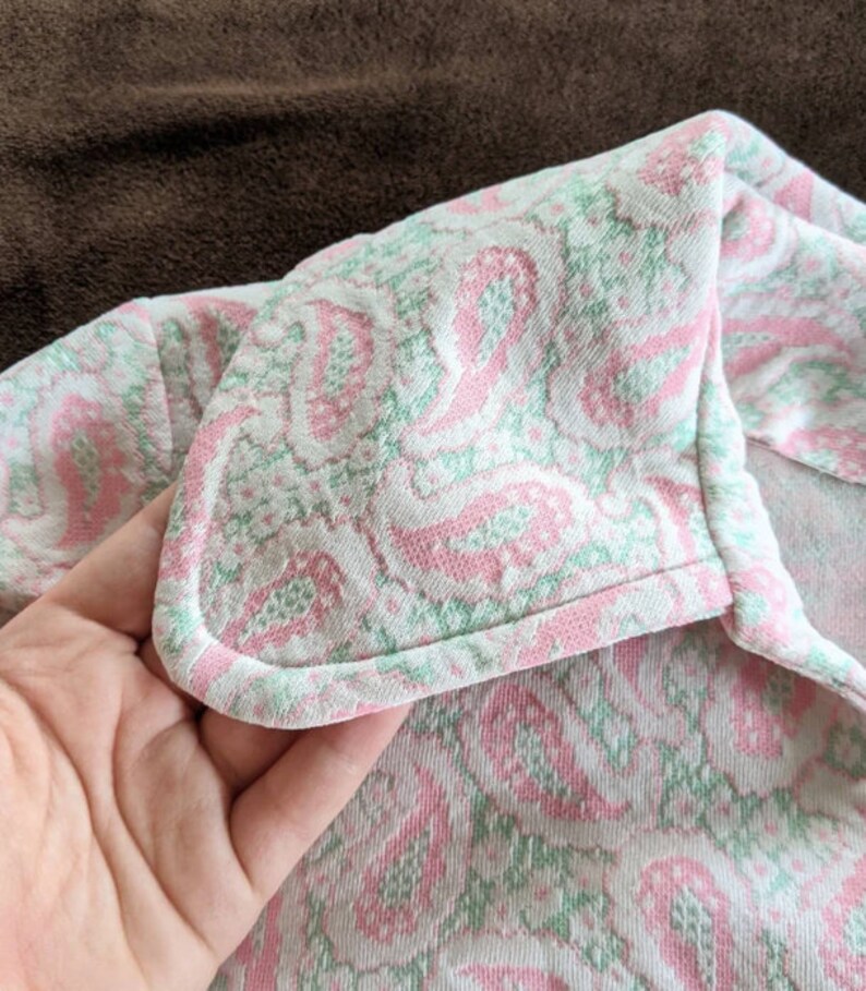 May include: Close-up of a pink and green patterned fabric with a white background. The fabric has a scalloped edge and is folded over.