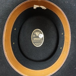 Puede incluir: El interior de un sombrero de fieltro negro con una banda de cuero marr&oacute;n. Una etiqueta dorada dice "SUPERCRAFT FUR FELT" con una etiqueta de tama&ntilde;o que dice "57 7".