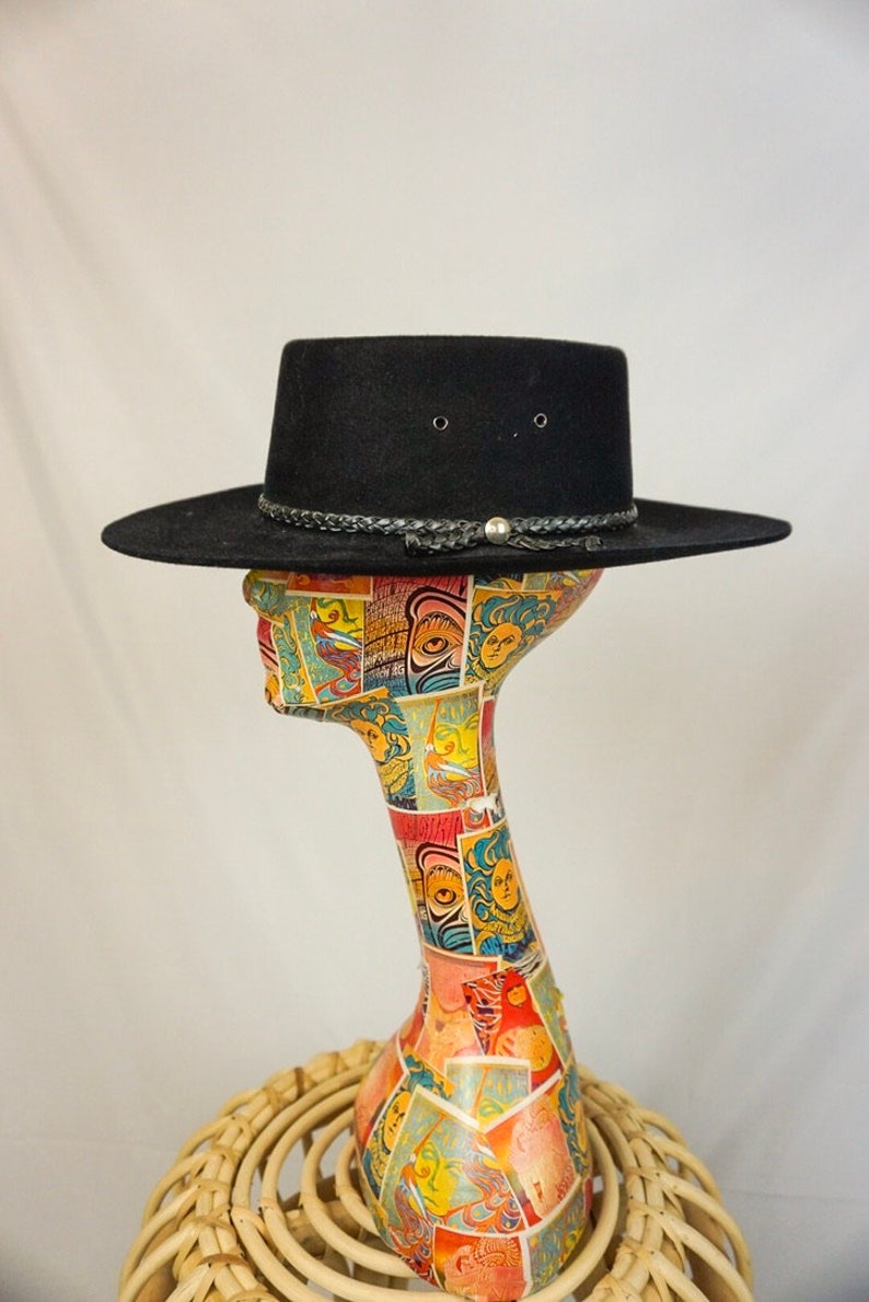 Puede incluir: Un sombrero vaquero de fieltro negro con una banda de cuero trenzada y una hebilla plateada. El sombrero est&aacute; sentado en una cabeza de maniqu&iacute;.