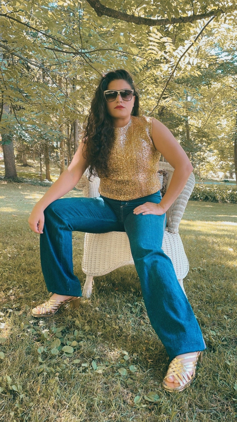 May include: A woman wearing a gold sequined sleeveless top, blue denim jeans, and gold sandals. She is sitting on a white wicker chair in a grassy area.