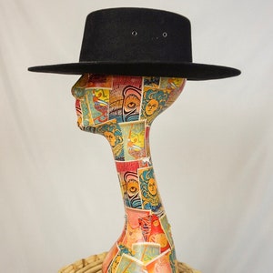 Puede incluir: Un sombrero fedora negro de ala ancha, sentado sobre una cabeza de maniqu&iacute; colorida. El maniqu&iacute; est&aacute; sobre un taburete de mimbre tejido.