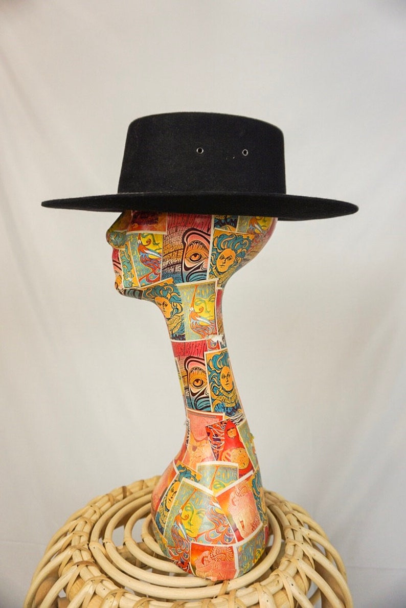 Puede incluir: Un sombrero fedora negro de ala ancha, sentado sobre una cabeza de maniqu&iacute; colorida. El maniqu&iacute; est&aacute; sobre un taburete de mimbre tejido.