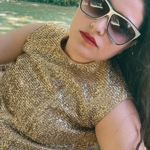 May include: A woman wearing a gold sparkly sleeveless top and sunglasses. She is sitting on a white wicker chair.