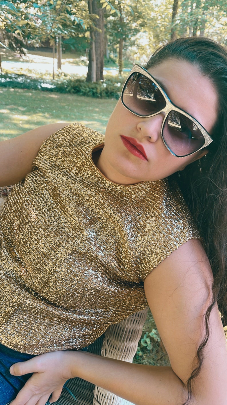 May include: A woman wearing a gold sparkly sleeveless top and sunglasses. She is sitting on a white wicker chair.