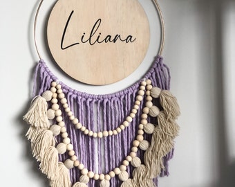 May include: A wooden circle with the name "Liliana" written in black script hangs from a gold hoop. The hoop is decorated with a lavender and cream macrame design with wooden beads.