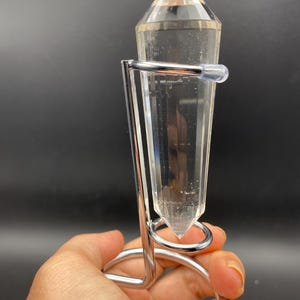 Op de afbeelding: Een heldere kwartskristallen slinger met een puntige punt, gehouden in een zilverkleurige metalen standaard. Het kristal is transparant en facetgeslepen, waardoor het licht reflecteert. De standaard heeft een cirkelvormige basis en een gebogen ontwerp. De achtergrond is donker.