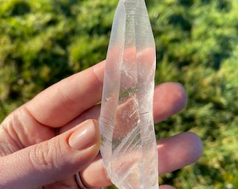 以下が含まれることがあります： 長く細い形をした透明な水晶ポイント。クリスタルは滑らかで磨かれており、わずかに湾曲しています。クリスタルは手で持たれており、緑の草の背景があります。