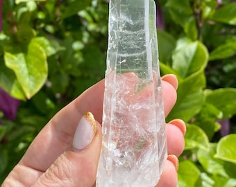 May include: A clear, elongated quartz crystal point with a natural, multifaceted structure. The stone is transparent with visible internal fractures and inclusions, tapering to a sharp, defined termination at the top and a rough, textured base.