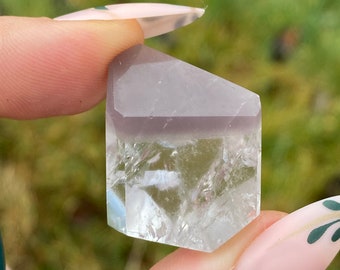 May include: A clear quartz crystal with a light purple top section. The crystal is held between two fingers, showcasing its geometric facets and translucent quality. The background is blurred green.