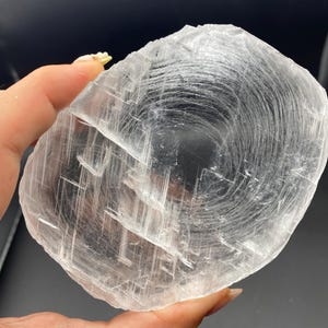 May include: A circular, translucent selenite crystal slab featuring a distinct, swirling fibrous pattern throughout its surface. The mineral displays a clear to white coloration with visible internal fractures and a layered, crystalline texture.