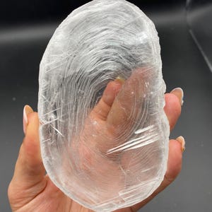 May include: A translucent, oval-shaped selenite crystal slab with a smooth, polished surface. The stone features visible internal fibrous striations and natural linear inclusions that create a layered, swirling pattern throughout the clear white mineral.