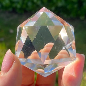 Puede incluir: Una pirámide de cristal transparente y facetada con un diseño geométrico. La pirámide se sostiene sobre un fondo verde, con la luz reflejada en las superficies del cristal. El objeto es transparente e incoloro.