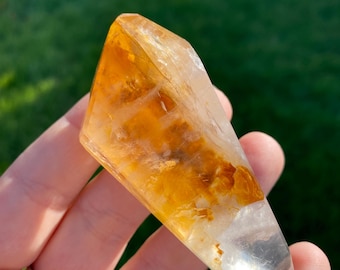 May include: A clear quartz crystal with a golden yellow inclusion. The crystal is shaped like a point and has a smooth, polished surface.