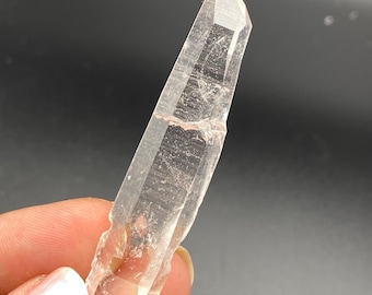 May include: A clear quartz crystal point with a natural, raw finish. The crystal is long and slender, with a pointed tip and a flat base. It is held in a hand for scale.