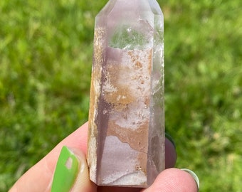 May include: A clear, faceted crystal point with a purple and brown color pattern. The crystal is held in a hand.