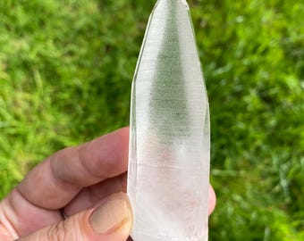 May include: A clear quartz crystal point, held against a background of green grass. The crystal is transparent with visible internal structures and a sharply defined tip. The natural light enhances its clarity and shape.