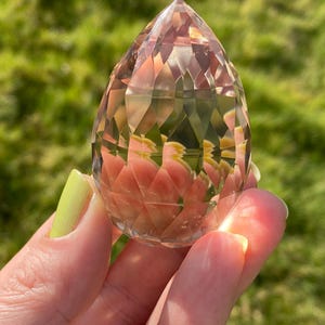May include: A clear, faceted crystal point in the shape of a teardrop. The crystal is held in a hand and has a slight pink hue.