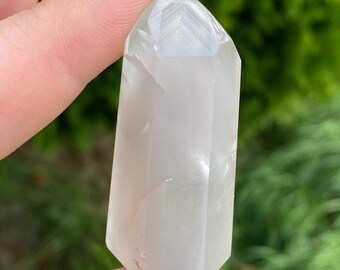 May include: A clear, white, faceted crystal point with a slightly cloudy appearance. The crystal is held in a hand with blue nail polish.
