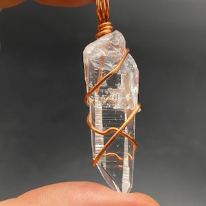 May include: A clear, elongated quartz crystal pendant wrapped in a thin, spiraling copper wire. The wire forms a small loop at the top for suspension, securing the raw, transparent stone which features natural internal fractures and facets.