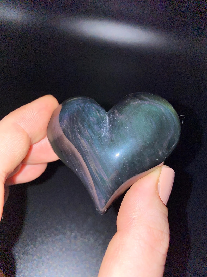 Puede incluir: Un cristal de obsidiana en forma de coraz&oacute;n con matices iridiscentes verdes y azules. El cristal est&aacute; pulido y liso.