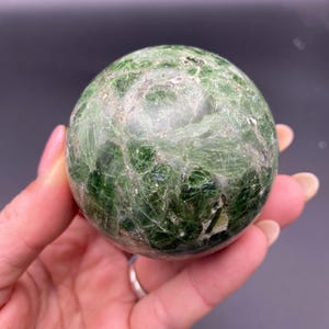 May include: A polished, spherical gemstone in shades of green. The orb has a textured surface with visible veining and inclusions. The sphere is held in a hand, showcasing its size and detail. Ideal for display or collection.