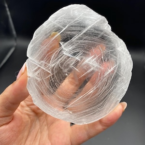 May include: A circular, translucent selenite crystal disc featuring a fibrous, layered texture with visible internal fractures and a clear, glass-like appearance. The mineral specimen displays a natural, radial pattern of fine, white, thread-like inclusions.