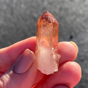 May include: A clear quartz crystal with a reddish-brown hue. The crystal is pointed at the top and has a natural, rough texture.