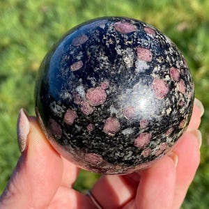 May include: A polished sphere of black granite with small, round, pinkish-red gemstones. The sphere is held in a hand against a blurred green background, highlighting the natural stone's unique pattern and colour variations.