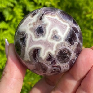 May include: A polished spherical stone featuring deep purple amethyst sections contrasted by intricate, branching white and light lavender quartz patterns. The surface is smooth and rounded, displaying a complex, natural geometric network of mineral inclusions.