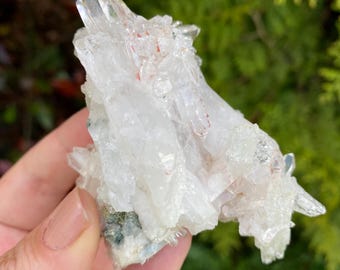 Può includere: Un gruppo di cristalli di quarzo trasparente con una consistenza naturale e ruvida. I cristalli hanno un aspetto traslucido con sfumature di inclusioni rosse e verdi. Il quarzo è tenuto in mano su uno sfondo di fogliame verde.