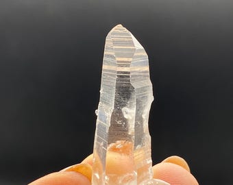 May include: A clear quartz crystal with a pointed tip and visible internal striations. The crystal is held against a dark background, showcasing its translucent quality and natural form. The crystal is a natural mineral specimen.