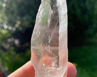 May include: A clear quartz crystal point with a sharp tip and a smooth, polished surface. The crystal is held in a hand, and the background is blurred.
