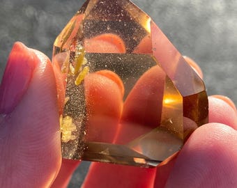 May include: A clear, faceted, golden citrine crystal point with inclusions. The crystal is held in a hand.