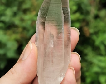 May include: A clear quartz crystal point with a pointed tip and a smooth, polished surface. The crystal is held in a hand, showcasing its size and shape.