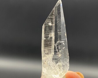 May include: A clear, elongated crystal with a sharp, pointed tip. The crystal has visible internal striations and a rough, textured base. The overall color is translucent with subtle variations, held by a person's fingers.