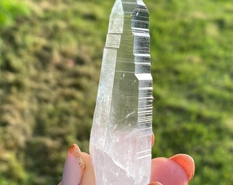 May include: A clear quartz crystal point with a faceted, elongated shape. The crystal is held against a backdrop of green grass, showcasing its transparency and natural lines. The crystal's tip is sharply defined.