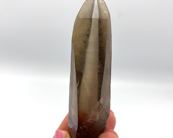 May include: A smoky quartz crystal, displaying a gradient of brown hues, from dark at the base to lighter at the pointed tip. The crystal's surface is smooth and polished, with natural inclusions. The crystal is held in a hand.