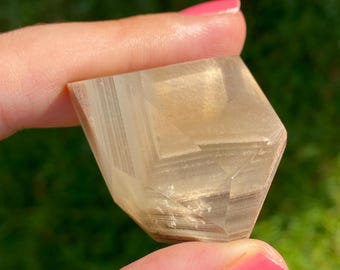 May include: A faceted, translucent crystal with a light brown hue. The crystal is held between two fingers against a blurred green background. The crystal's geometric shape is visible.