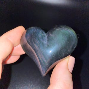 Puede incluir: Un cristal de obsidiana en forma de coraz&oacute;n con matices iridiscentes verdes y azules. El cristal est&aacute; pulido y liso.